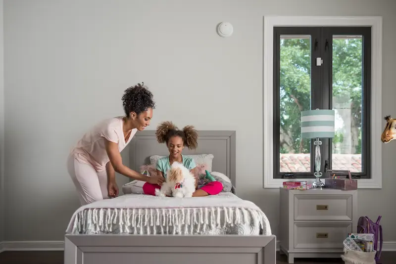 A woman and a young girl pet a small white dog on a bed in a room equipped with a wall-mounted smoke detector and a window sensor.